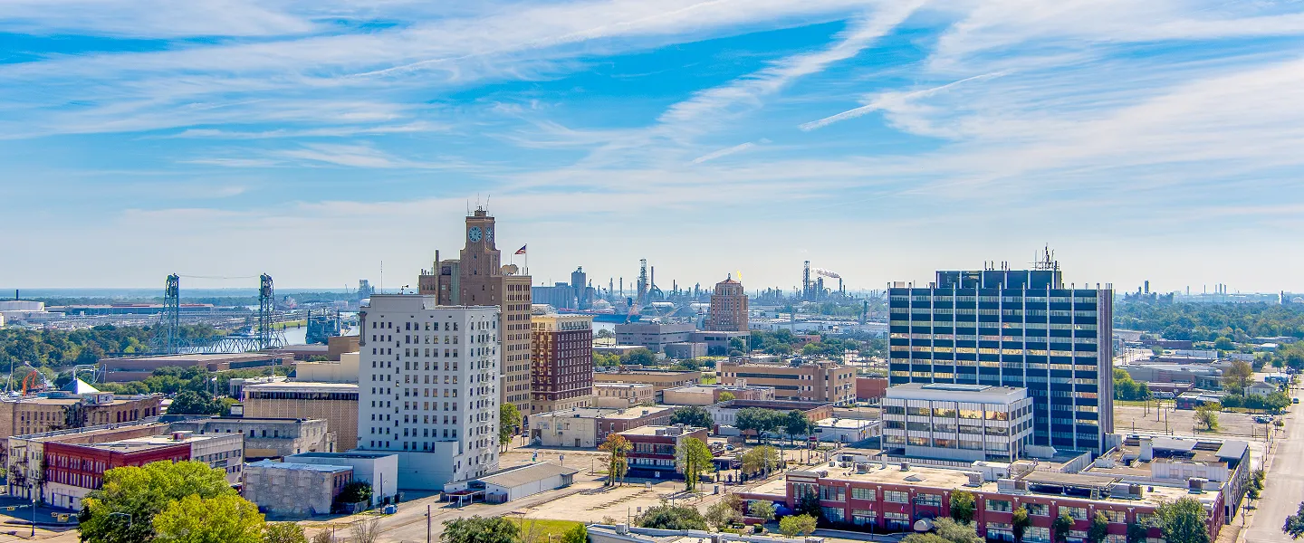 Panoramic cityscape featuring mid-rise buildings, industrial structures in the background, and a partly cloudy blue sky.