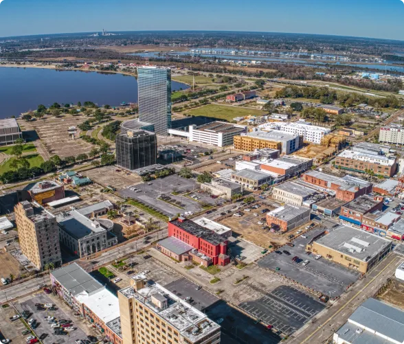 Aerial view of urban downtown area with mid-rise buildings, parking lots, and a river in the background.
