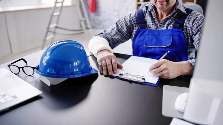 Engineer in blue overalls writing on clipboard next to blue safety helmet and glasses on desk.