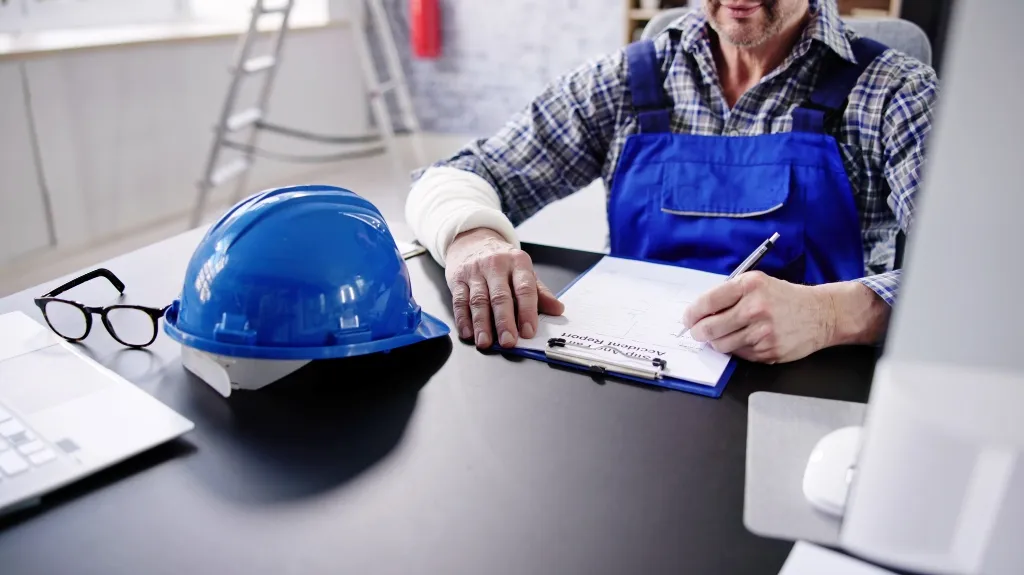 Engineer in blue overalls writing on clipboard next to blue safety helmet and glasses on desk.