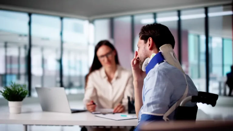 Man with neck brace sitting in wheelchair consulting with female professional in modern office.
