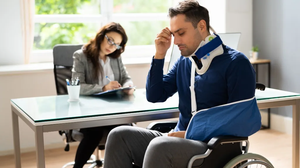Man with neck brace and arm sling sitting in wheelchair, woman in background writing at desk.