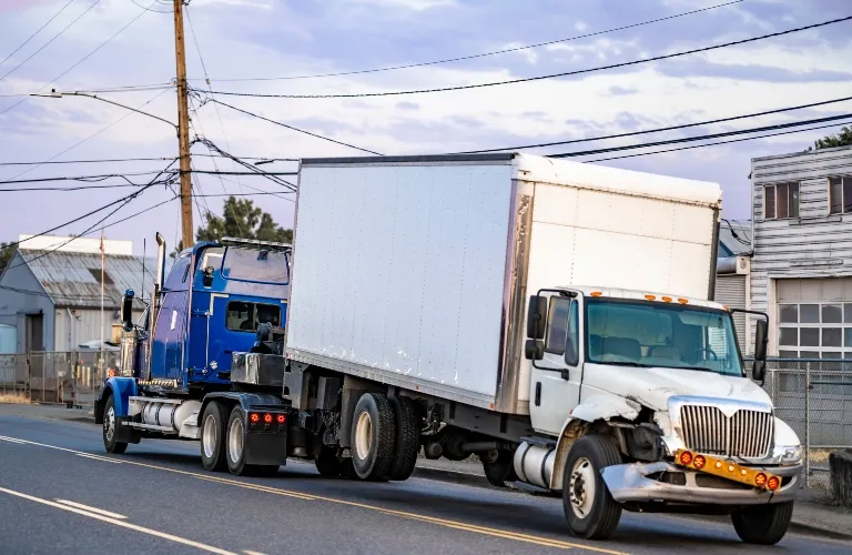 White box truck with front-end damage being towed by a blue semi-truck on urban road.