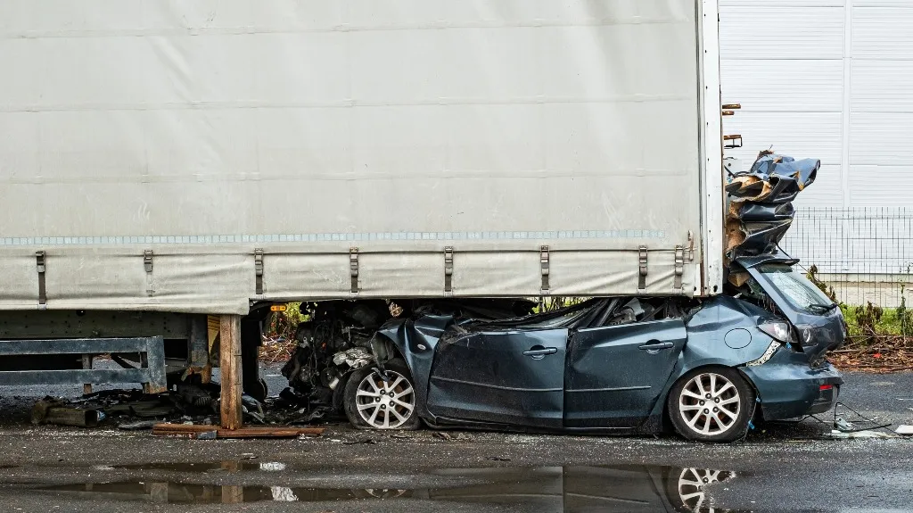 Dark blue sedan severely crushed underneath the rear of a large white truck trailer.